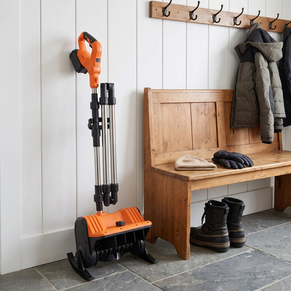 A tidy mudroom corner with an orange-and-black cordless snow shovel with its shaft folded standing upright against a white wall, a wooden bench at the right with plain winter boots, a beanie and gloves, and coat hooks above