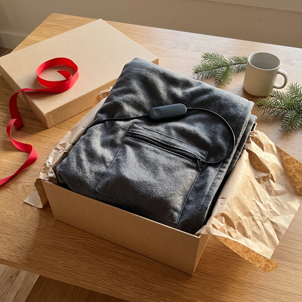 A plain kraft-paper gift box with its lid off and a red ribbon on a wooden table, the folded graphite-grey blanket lifted half out of crinkled brown paper with its pocket corner and a small black controller in front, a mug and pine sprigs at the side